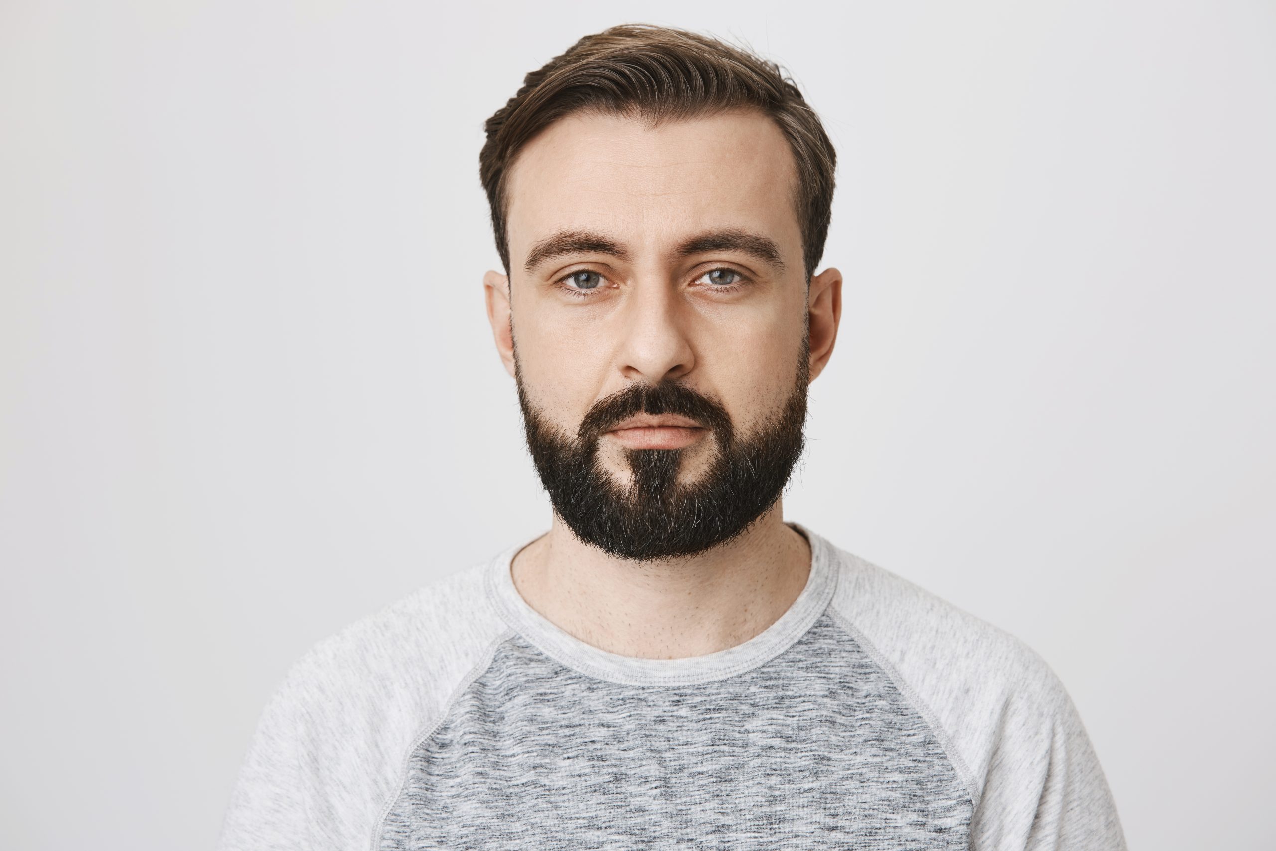Calm interesting bearded guy in trendy shirt with normal face expression, looking at camera while standing against gray background. Man takes photo for passport at local studio