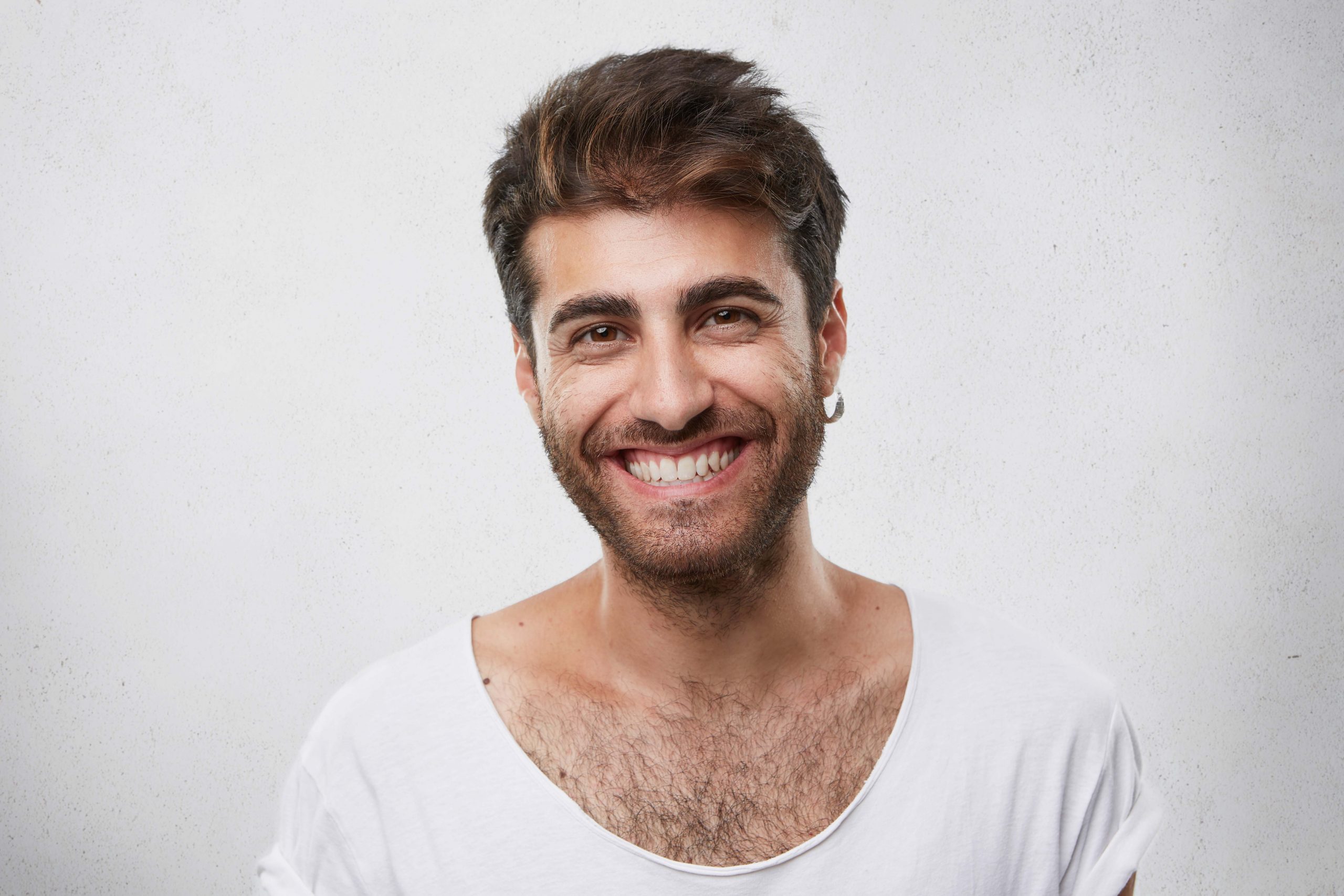 Positive bearded guy with dark warm eyes having earring in ear dressed casually having pleasant smile showing his perfect white teeth having good mood after party and meeting with his friends