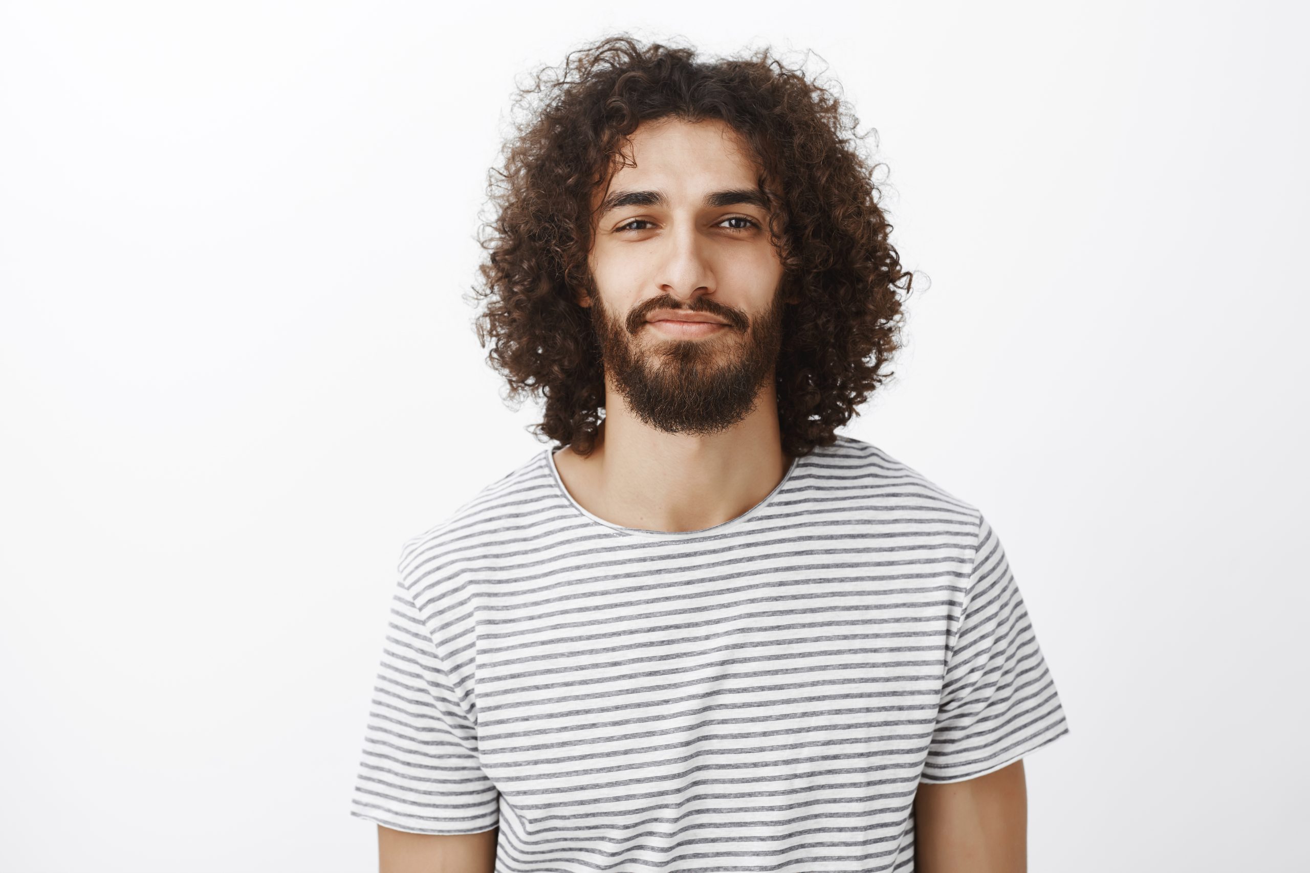 Portrait of handsome confident creative boyfriend with dark curly hair, standing with slight smile and self-assured expression, ready and in right mood to invite girlfriend to dance over gray wall