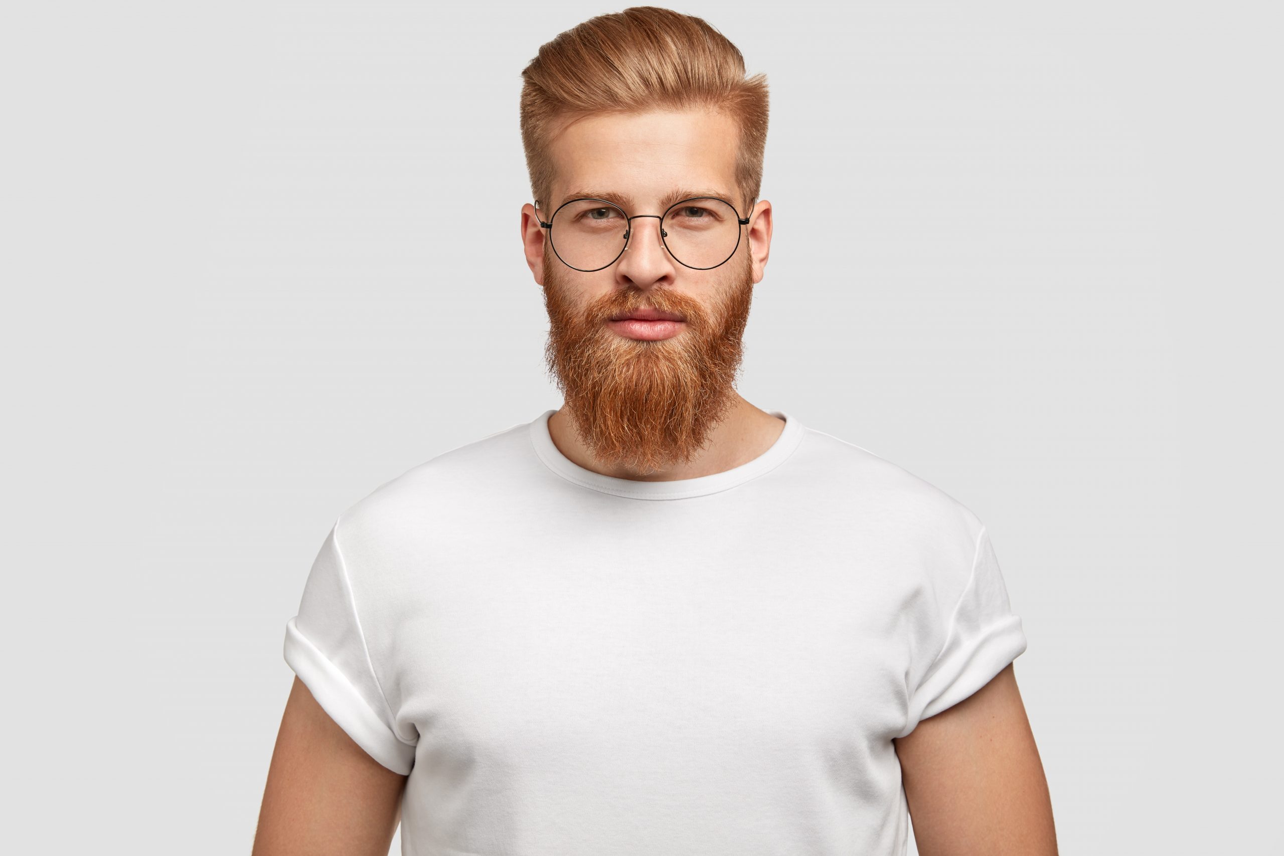 Portrait of handsome stylish male with trendy hairdo, looks seriously at camera, thinks about something, wears spectacles and casual t shirt, isolated over white background. Masculinity concept