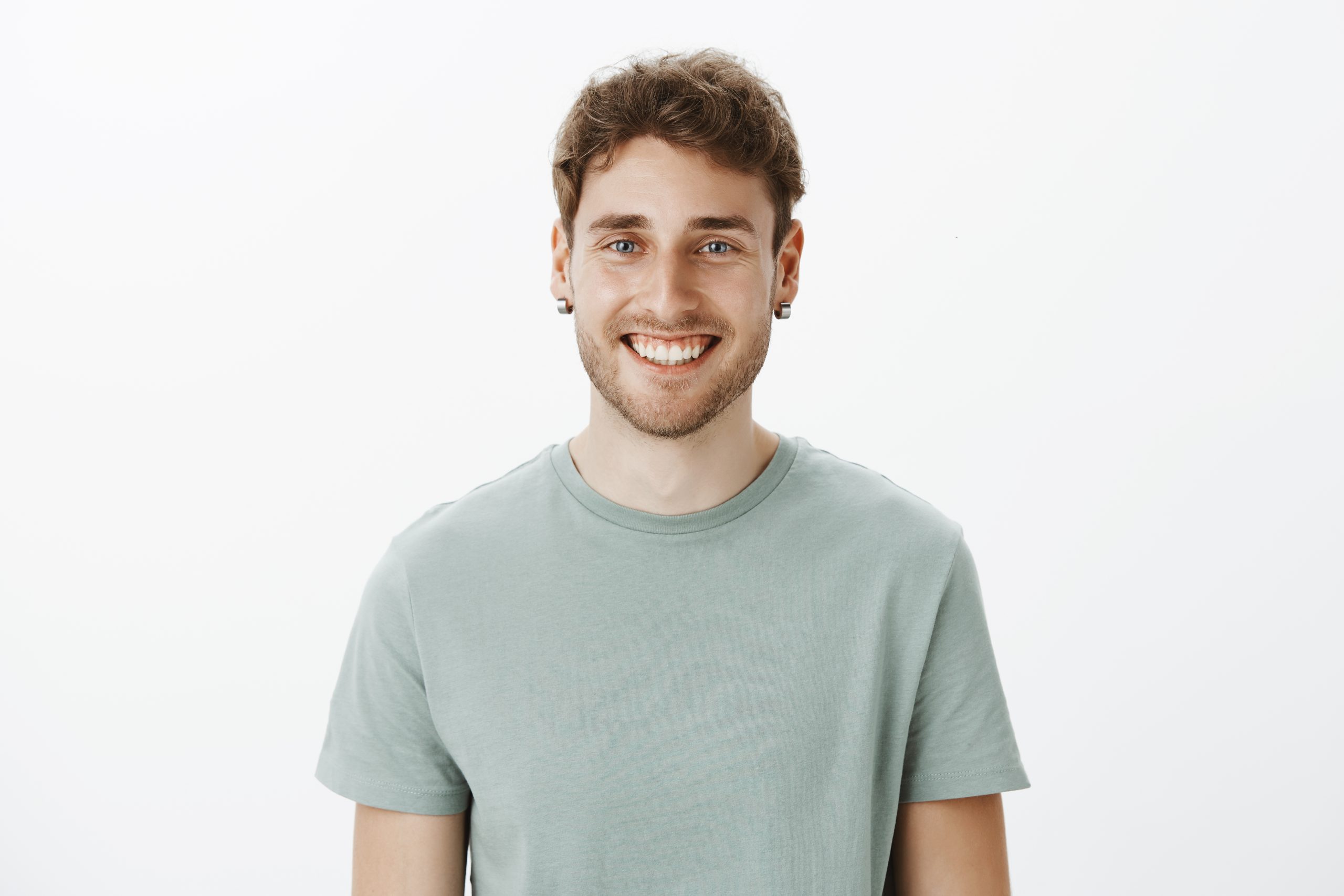 Portrait of satisfied happy Caucasian man in earrings and t-shirt, smiling broadly and gazing pleased at camera, talking casually with good friend, enjoying spending time with close people