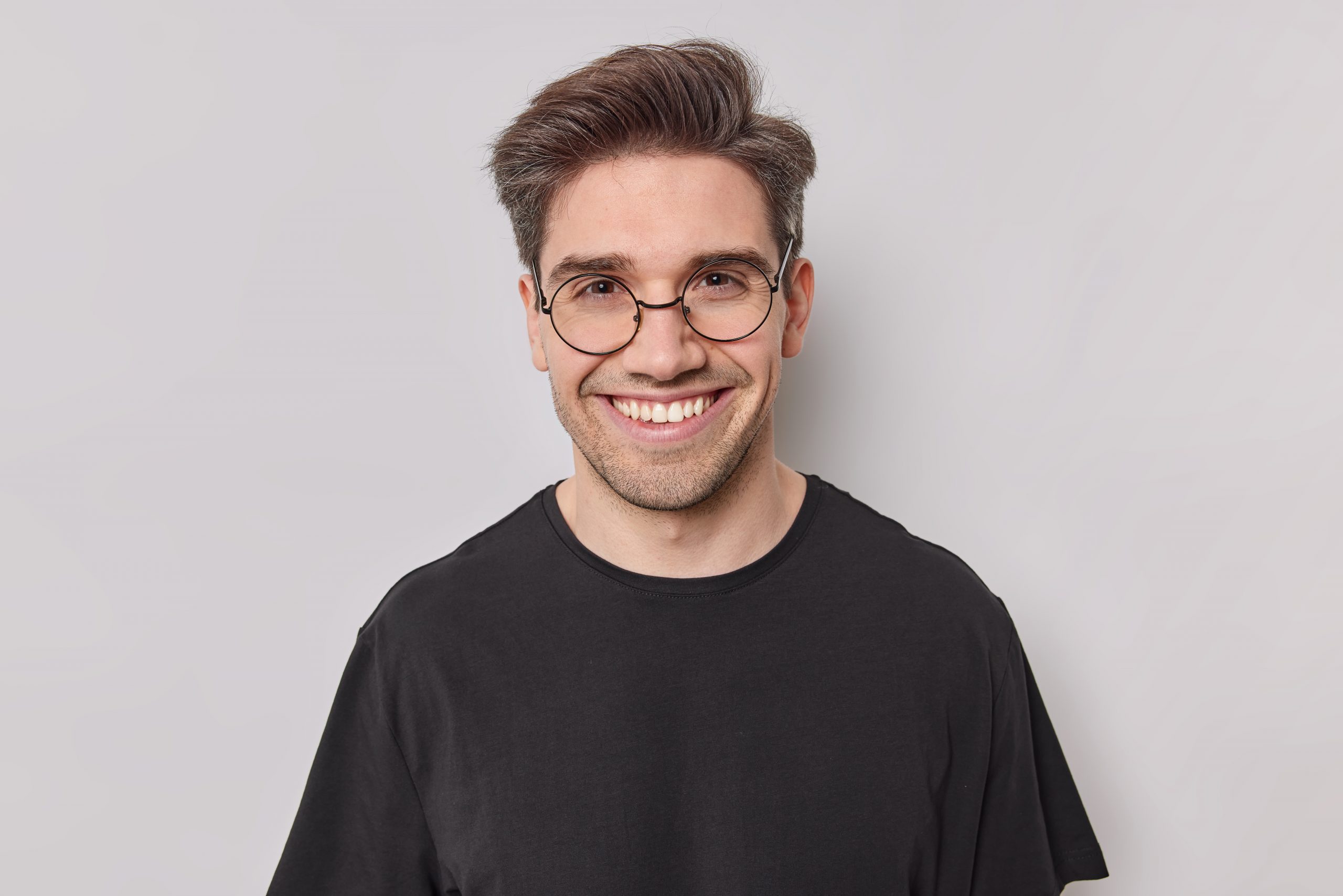 Portrait of happy man with dark hair smiles with white teeth looks confident at camera wears round transparent glasses black t shirt poses at studio against white background. Positive emotions